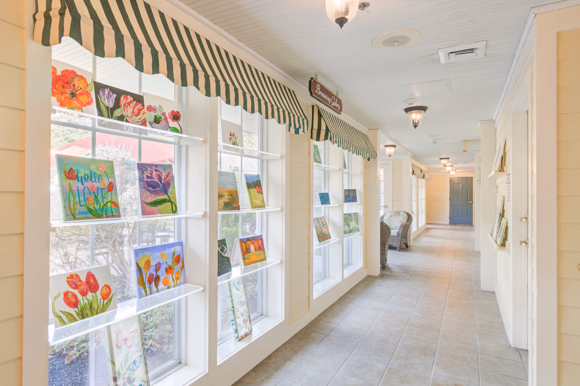 Sunlit interior corridor with large display windows showing colorful paintings, striped awnings, tiled floor, and seating.
