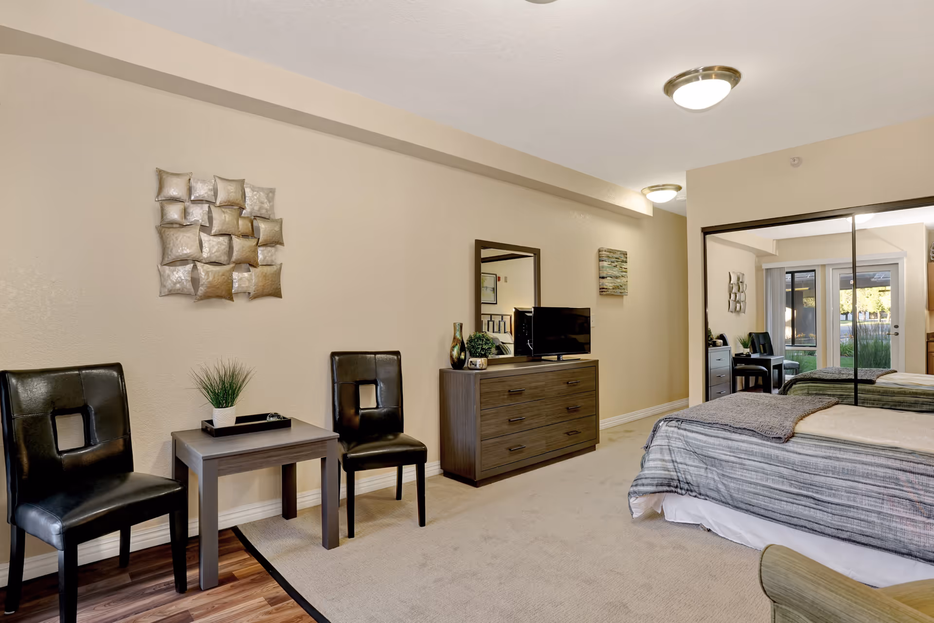A well-lit bedroom featuring a bed with a striped comforter, a dresser with a TV and decorative items, two black chairs with a small table between them, wall art, and a large mirrored closet reflecting the room and a glass door leading outside.