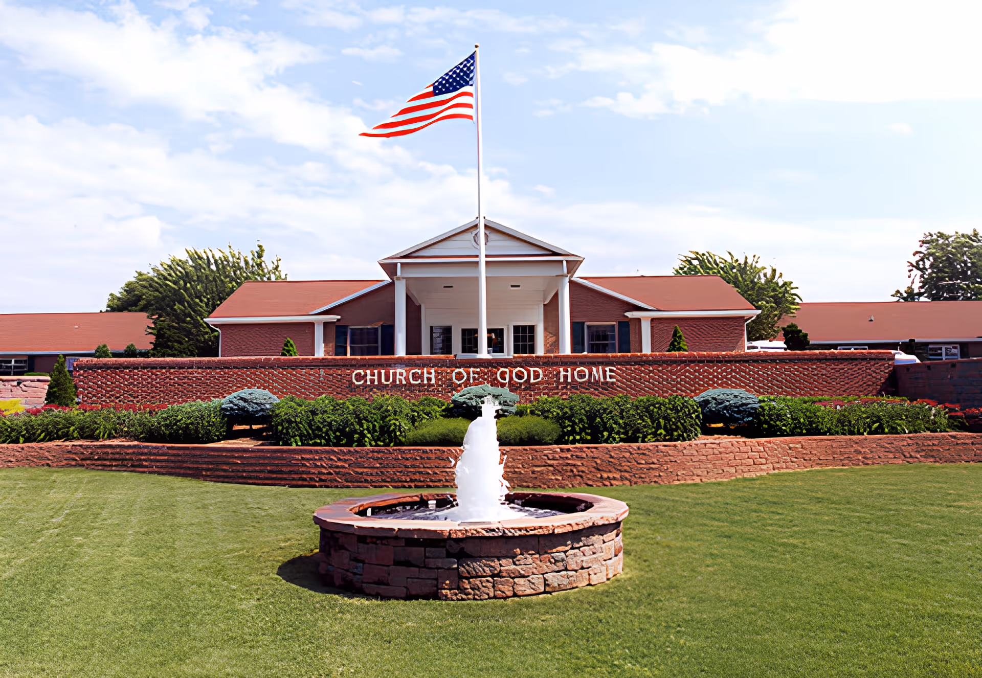 Front exterior view of the Church of God Home building with a red roof, brick facade, an American flag on a flagpole, a water fountain in the foreground, and well-maintained green lawn and shrubs.