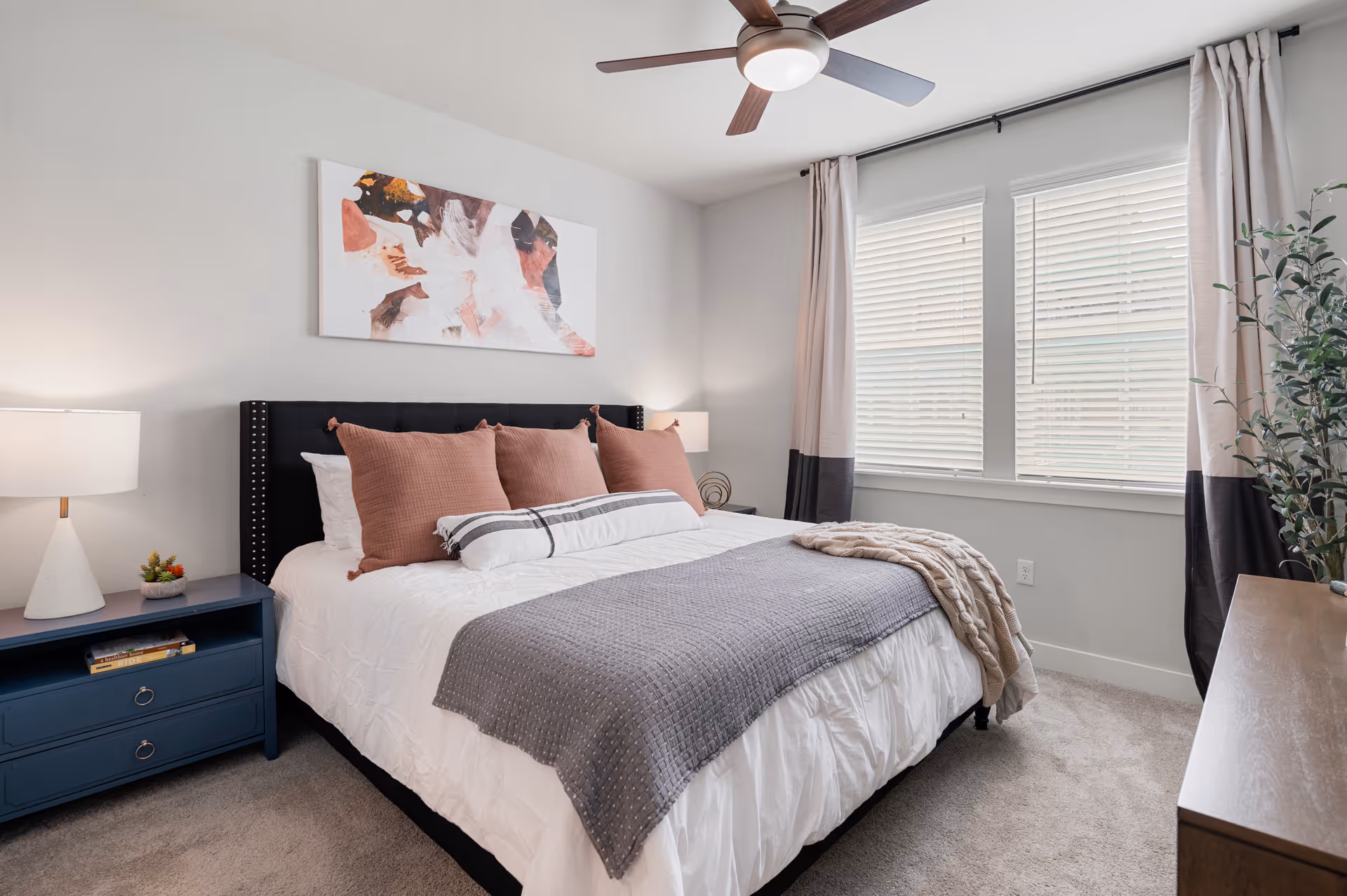 Bright modern bedroom with a made bed, decorative pillows and throw, nightstand with lamp, wall art, ceiling fan, and windows with blinds.