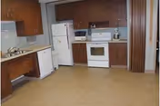 A kitchen area with wooden cabinets, a white refrigerator, a white stove with oven, a microwave, and a dishwasher. The floor is covered with beige linoleum, and there is a sink with a faucet on the left side.