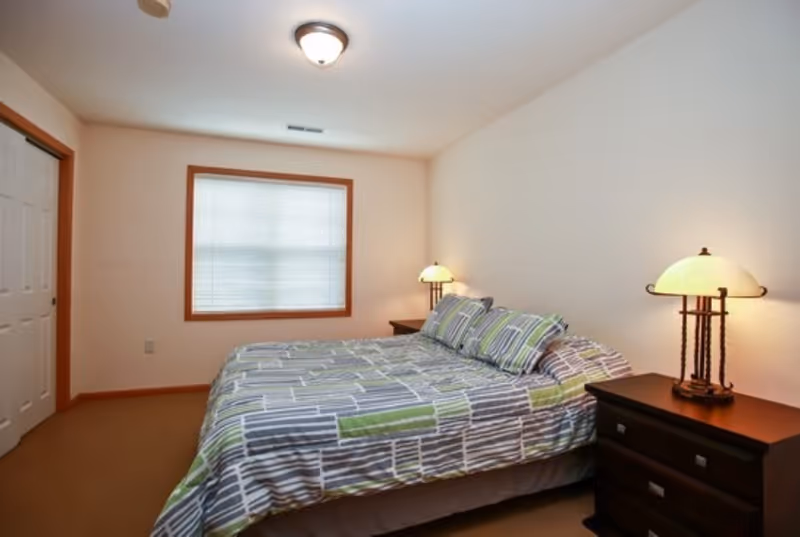 A bedroom with a bed covered in a striped bedspread in shades of gray, green, and white. There are two matching pillows on the bed. On each side of the bed, there is a dark wooden nightstand with a lamp featuring a round, cream-colored shade. The room has beige walls, a window with closed blinds, a ceiling light fixture, and a closet with sliding white doors.