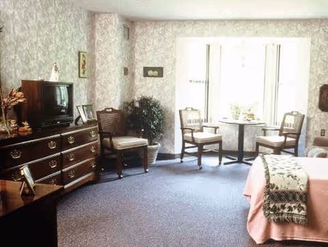 Sunlit senior living bedroom with a bed, dresser and TV, and a seating area by a large window.