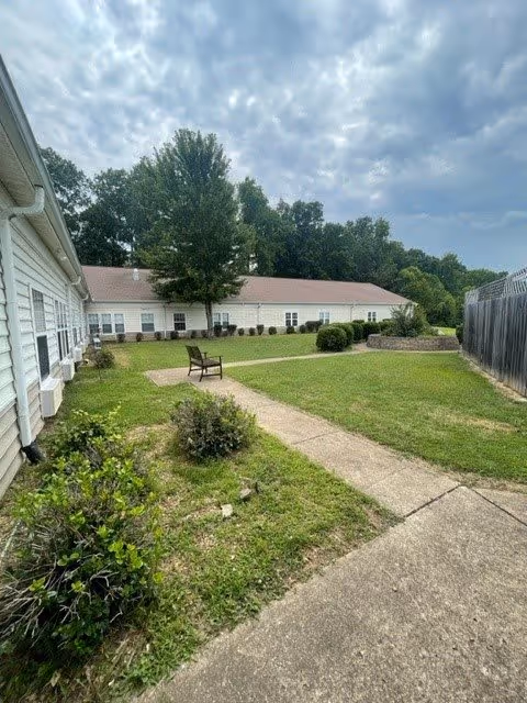 Outdoor courtyard area of a senior living facility with a concrete pathway, green grass, bushes, a single chair, and a large tree. The building with white siding and multiple windows surrounds the courtyard. The sky is partly cloudy.