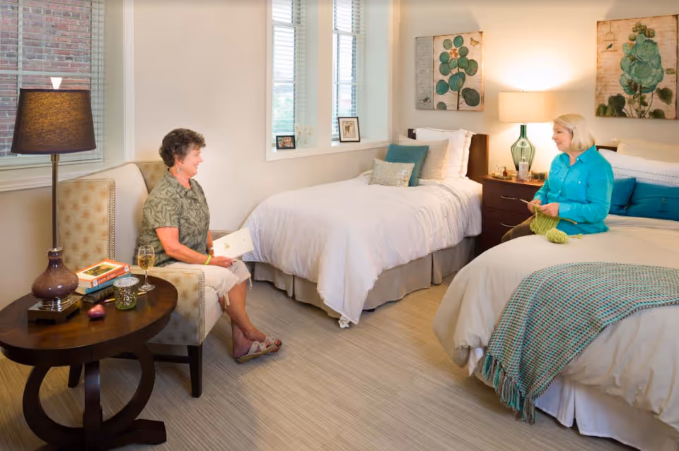 Two elderly women sitting and chatting in a well-lit bedroom with two beds, one woman sitting on an armchair holding a book and the other sitting on a bed knitting. The room has a side table with a lamp, books, and a glass of wine, with decorative paintings on the wall.