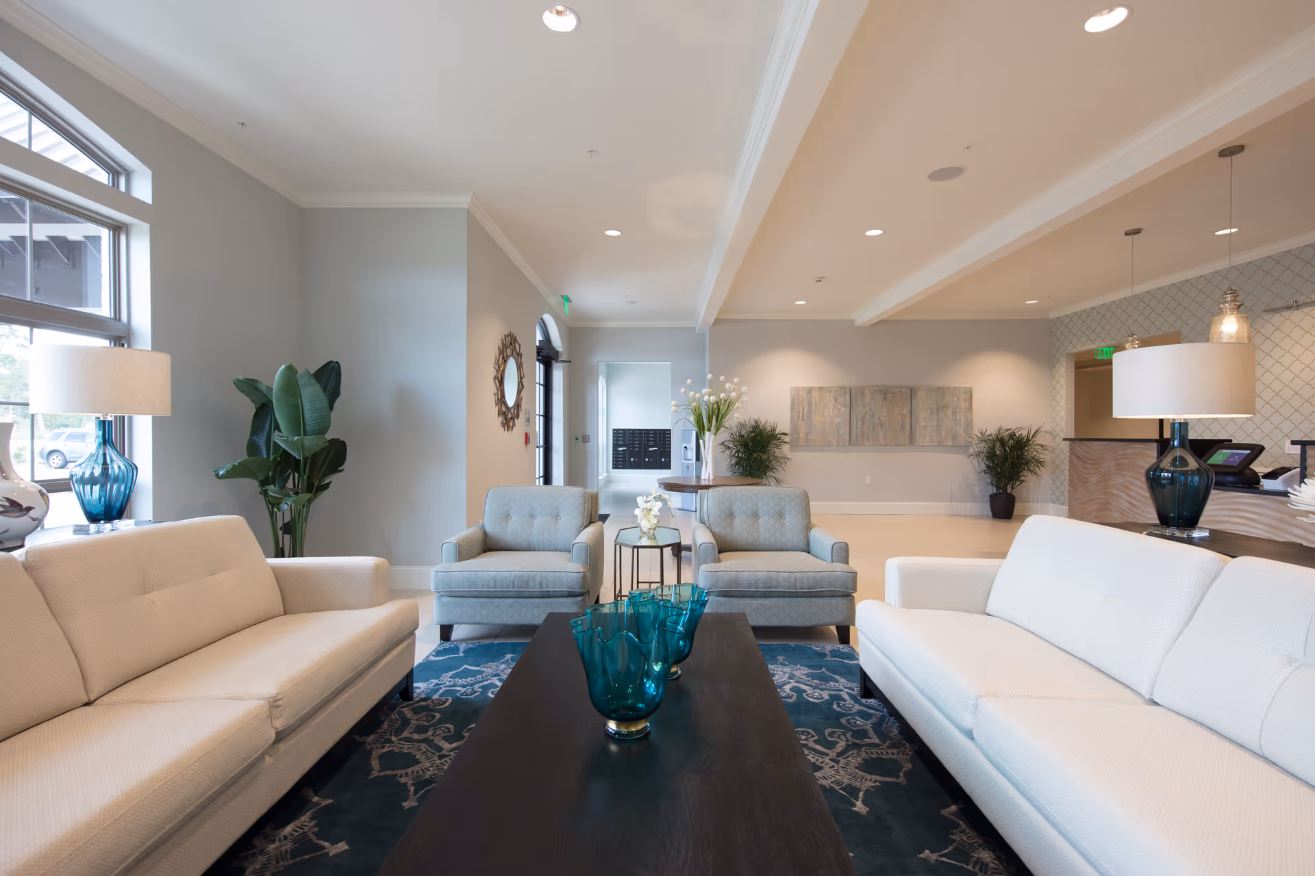 A bright and modern living room area with two white sofas facing each other, two light blue armchairs, a dark wooden coffee table with blue glass vases, a patterned blue rug, large windows letting in natural light, and decorative plants and lamps.