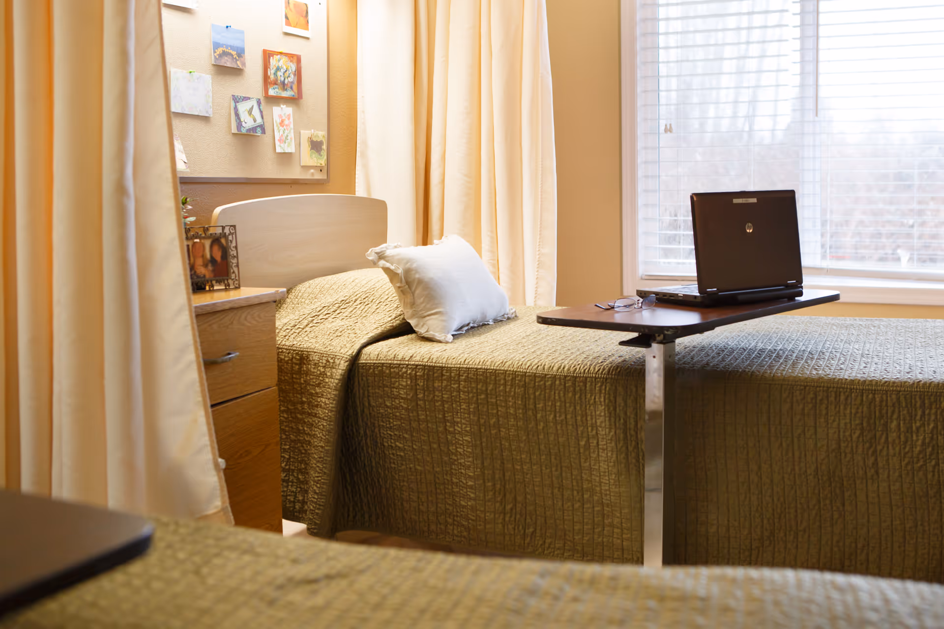 A small bedroom in a senior living facility with a single bed covered in a green quilt. A white pillow rests on the bed. Next to the bed is a wooden nightstand with a framed photo and a bulletin board on the wall displaying various colorful notes and pictures. A laptop and a pair of glasses are placed on an overbed table. The room is softly lit by natural light coming through a window with white blinds.