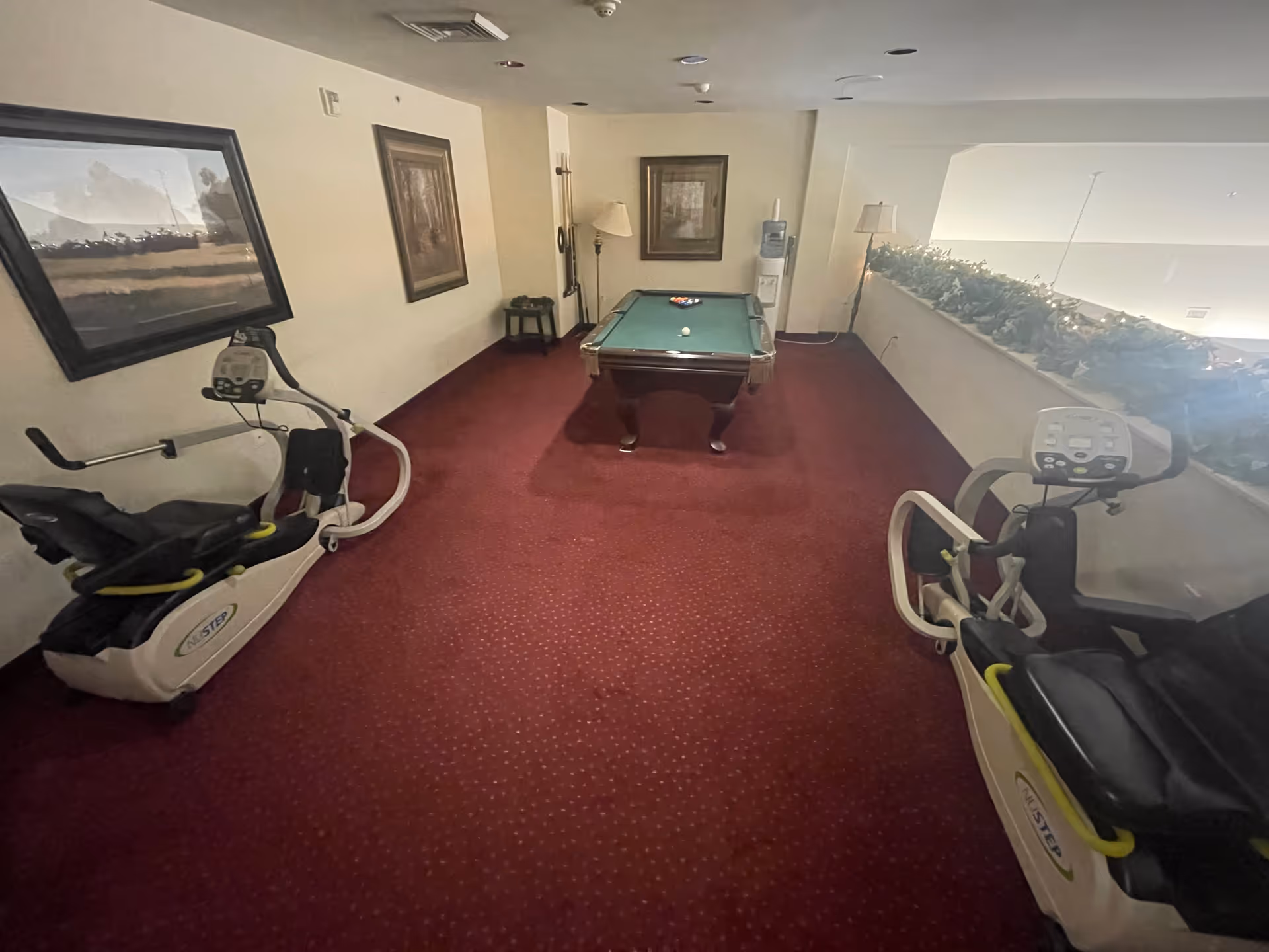A recreational room with a pool table in the center, two exercise bikes on either side, red carpeted floor, framed paintings on the walls, a water cooler, and a floor lamp near the back wall.
