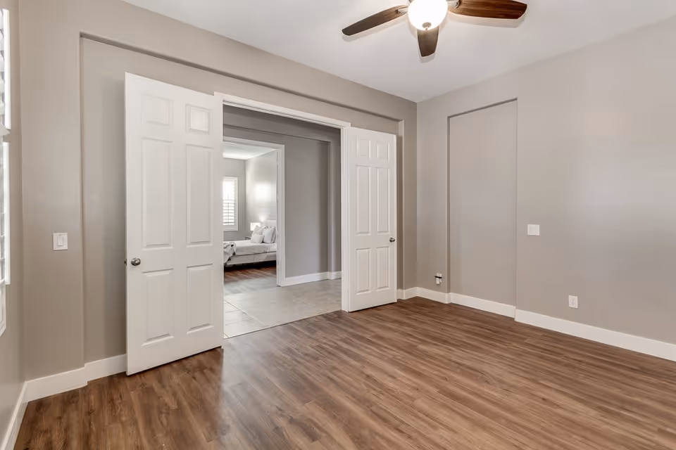 Empty interior room with wood-look flooring, a ceiling fan, and open double doors revealing a bedroom.