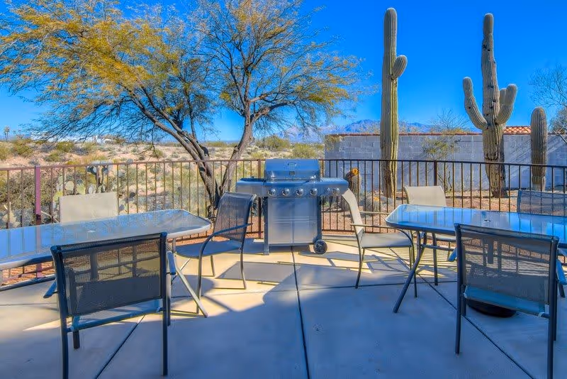 Outdoor patio area with metal tables and chairs arranged around a stainless steel barbecue grill. The patio is enclosed by a metal railing and surrounded by desert landscaping including cacti and a tree, with mountains visible in the background under a clear blue sky.