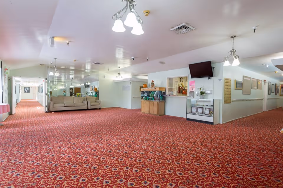 Interior view of a senior living facility hallway with red patterned carpet, beige sofas, a reception window, a mounted TV, display cases, and multiple light fixtures on the ceiling. The hallway extends in two directions with framed pictures on the walls.