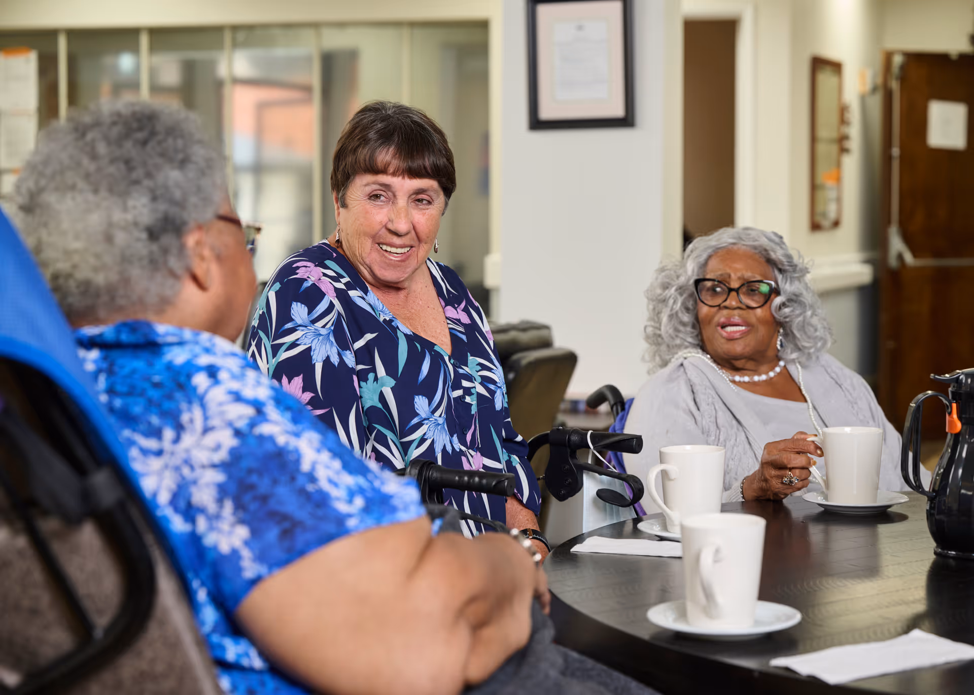 Three elderly women sitting around a table in a common area, engaging in conversation. Two of the women are in wheelchairs, and there are white coffee mugs and a coffee pot on the table. The setting appears to be a cozy, well-lit room with framed pictures on the walls.