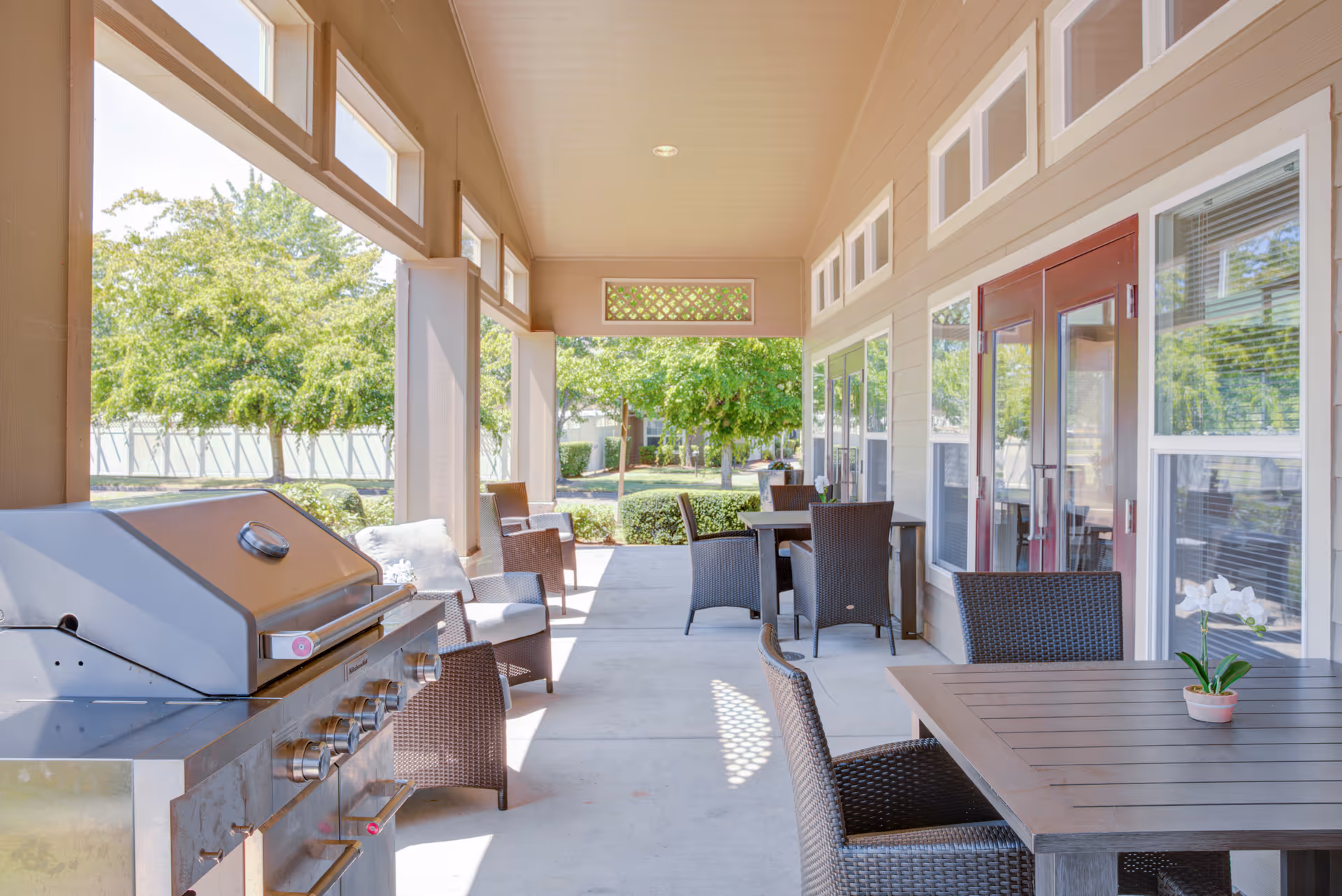 Covered outdoor patio area with a stainless steel grill on the left, several wicker chairs with cushions, and tables with potted plants. The patio overlooks a green garden with trees and bushes, and there are large windows and red double doors on the right side.