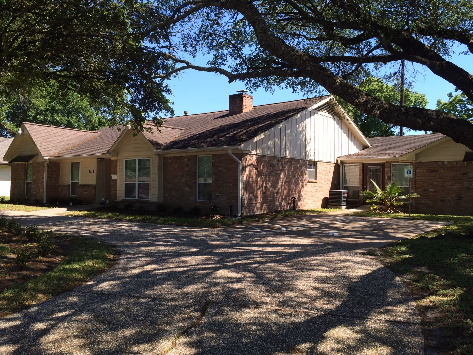 Single-story brick assisted living building with a circular driveway shaded by large oak trees.