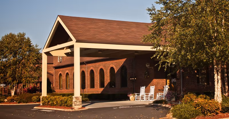 Brick retirement village building with a covered entrance and white rocking chairs outside.