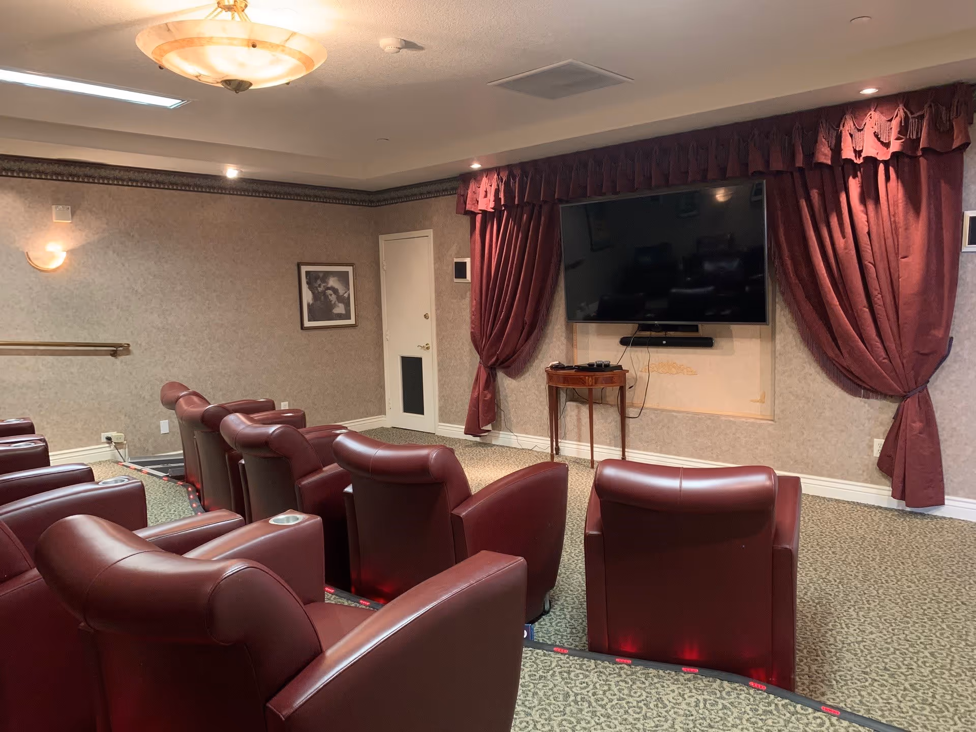A small theater room with rows of red leather recliner chairs facing a large flat-screen TV mounted on the wall. The room has beige walls, a patterned carpet, maroon curtains framing the TV, and soft overhead lighting.