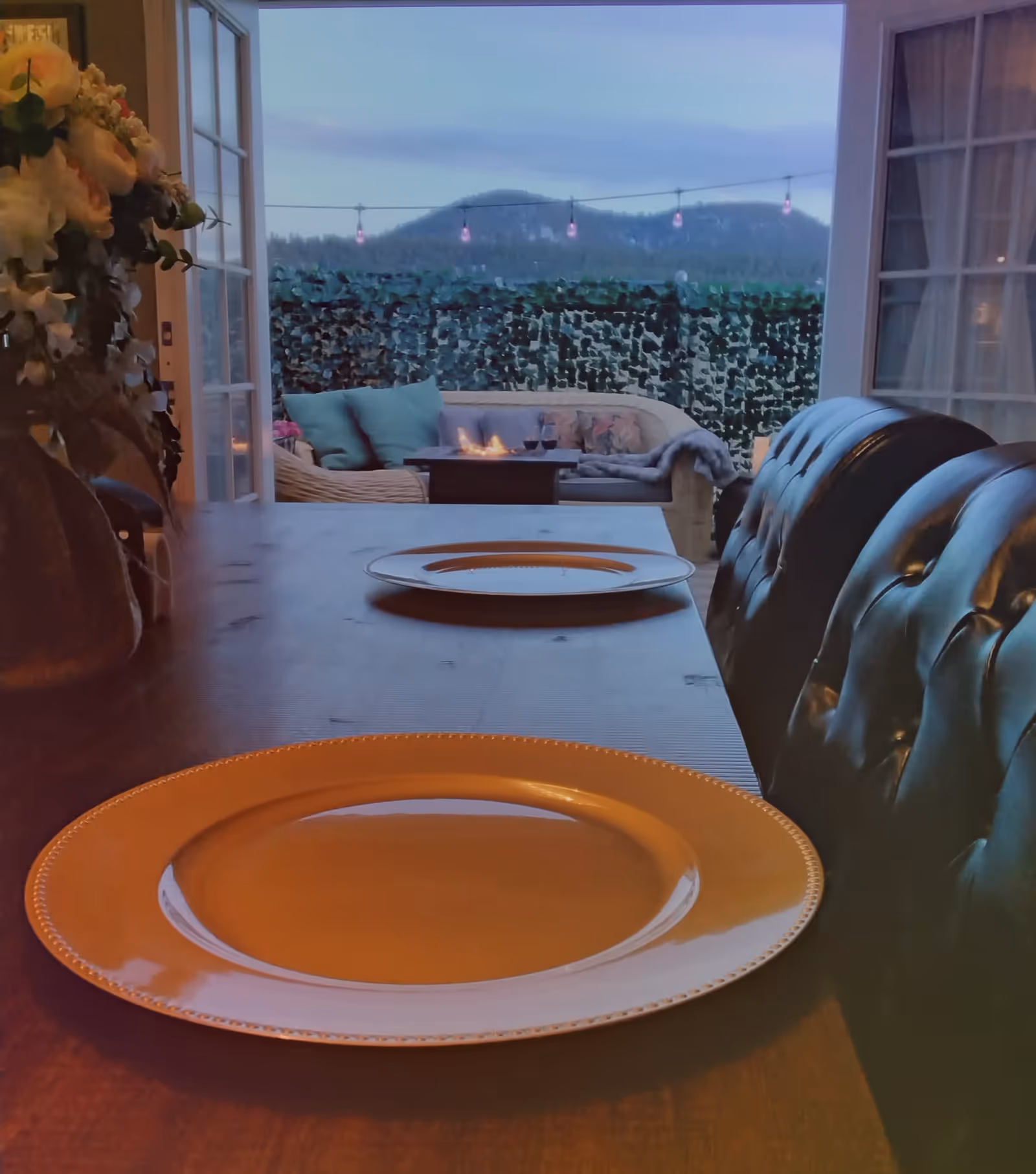 View of a dining table with two empty plates and leather chairs, looking out through open doors to an outdoor seating area with a fire pit, cushions, string lights, and a backdrop of hills under a twilight sky.