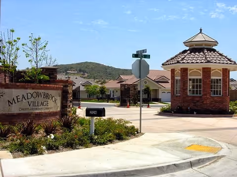 Entrance to Meadowbrook Village retirement community showing a brick sign, guardhouse, mailbox and homes with hills in the background.