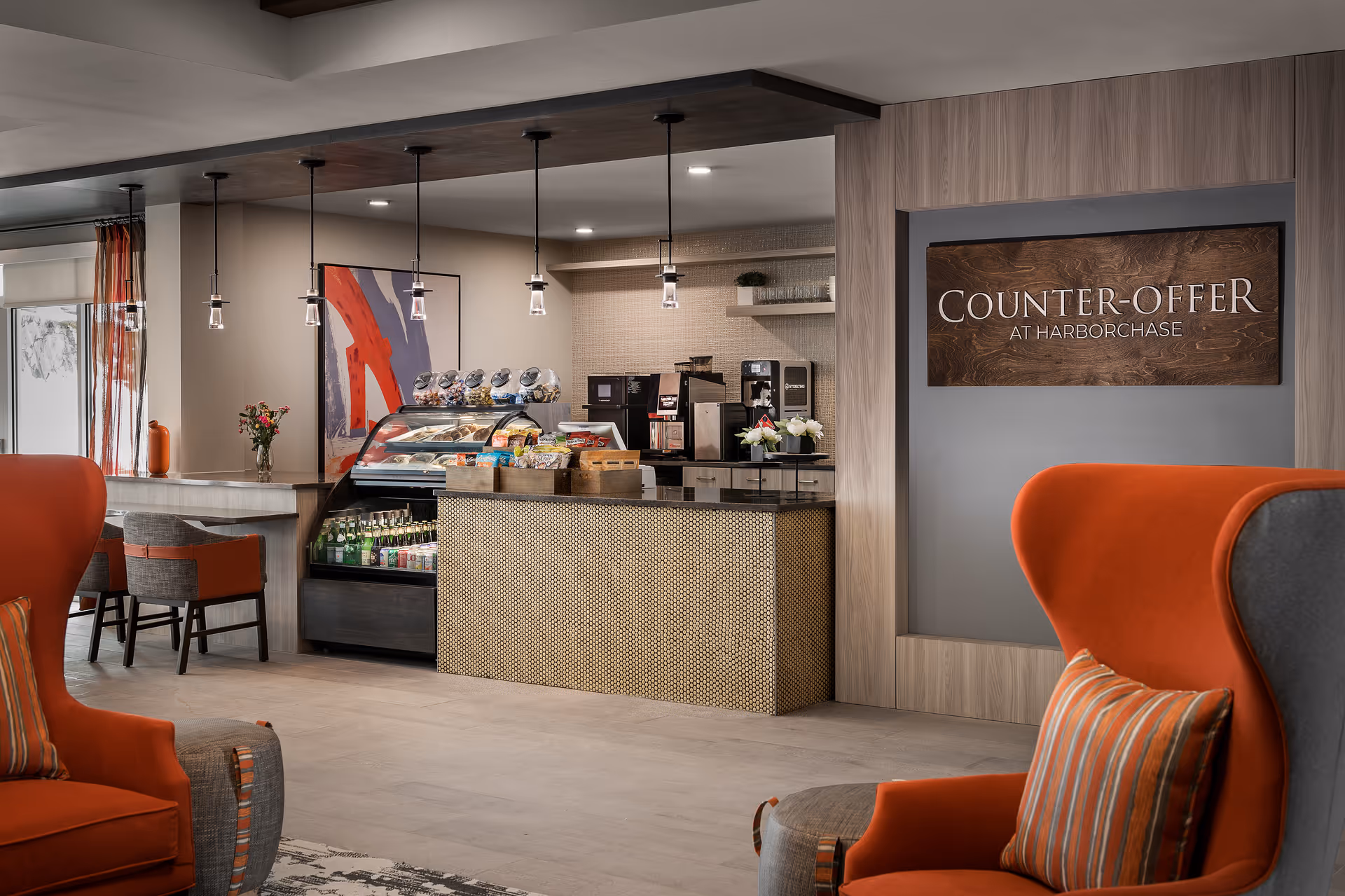 Interior view of a modern cafe area named Counter-Offer at HarborChase, featuring a counter with a display case of snacks and beverages, coffee machines, and seating with orange and gray armchairs and chairs. Pendant lights hang from the ceiling, and there is a colorful abstract painting on the wall.