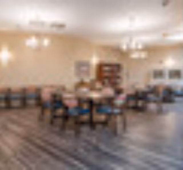 A spacious dining room with multiple round tables and chairs arranged neatly. The room has wooden flooring, beige walls, and is well-lit with ceiling lights and wall sconces. There is a wooden cabinet against the far wall and framed pictures hanging on the walls.