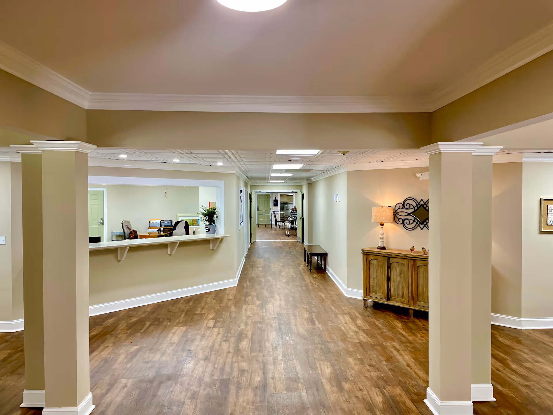 Interior hallway of a senior living facility with wooden flooring, beige walls, and white crown molding. On the left side, there is a reception window with chairs and office supplies visible behind it. On the right side, there is a wooden cabinet with a lamp and decorative wall art above it. The hallway extends into a room with tables and chairs in the background.