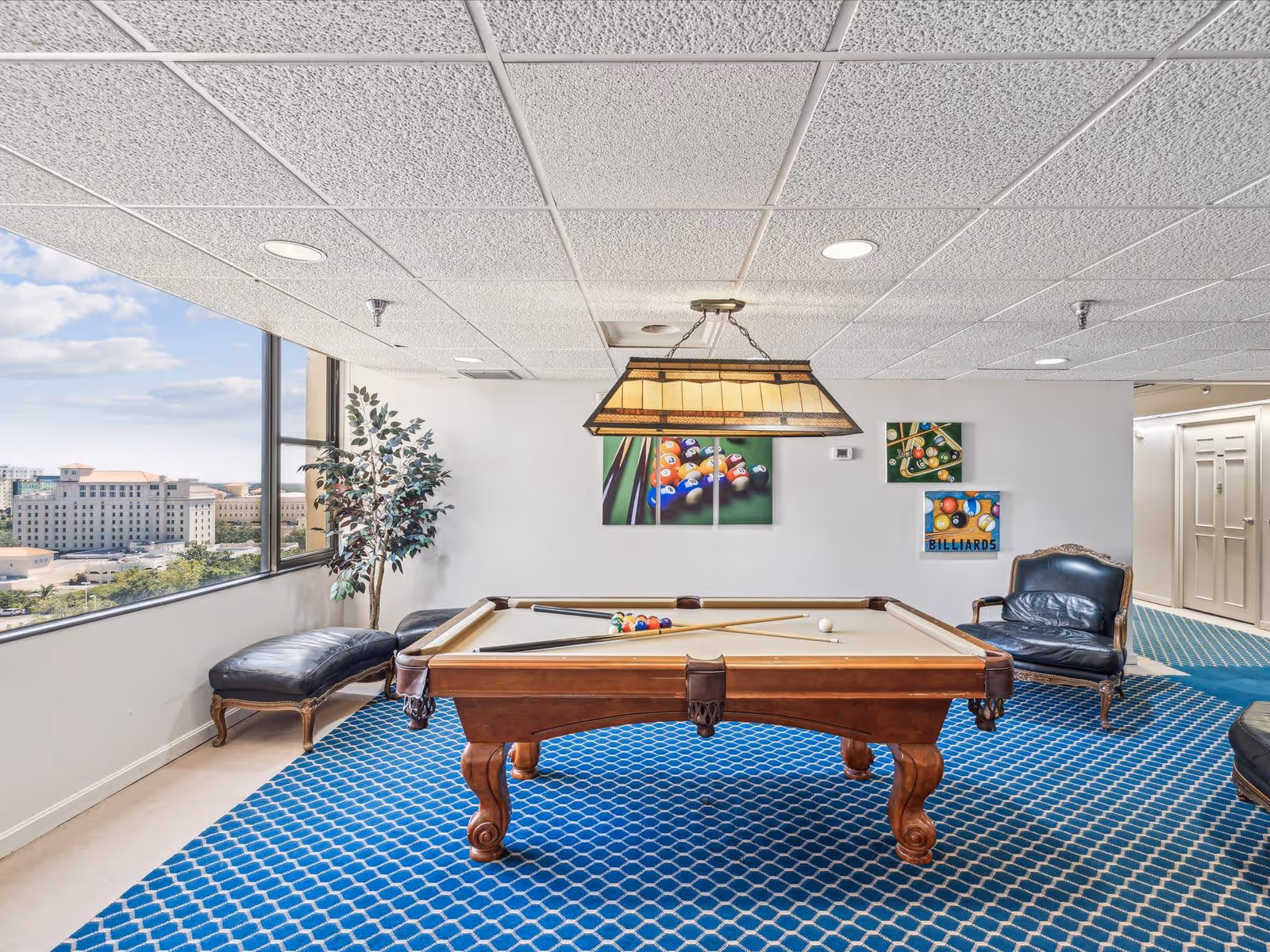 A bright game room with a pool table in the center, two pool cues resting on it, and billiard balls arranged on the table. The room has a blue patterned carpet, a large window showing a cityscape outside, a potted plant, a black leather chair, and three billiards-themed paintings on the white wall. A rectangular hanging light fixture is above the pool table.