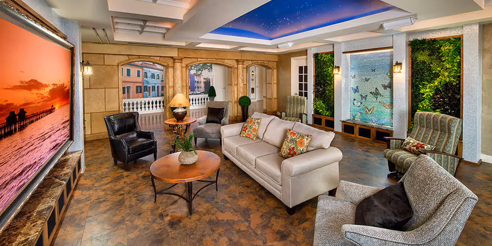 A spacious and elegant senior living lounge area with a beige sofa adorned with floral pillows, several armchairs, a round wooden coffee table with a plant, and a large wall-mounted screen displaying a sunset over the ocean. The room features decorative columns, a ceiling painted to resemble a starry sky, and vertical green plant walls flanking a butterfly-themed water feature.