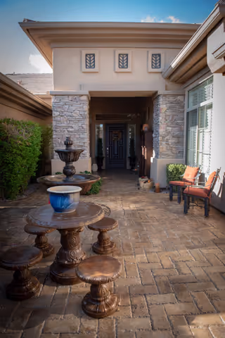 Outdoor patio area with a stone table and stools, a water fountain, and two cushioned chairs near a window. The entrance to the building is visible in the background with stone pillars and decorative windows above the doorway.