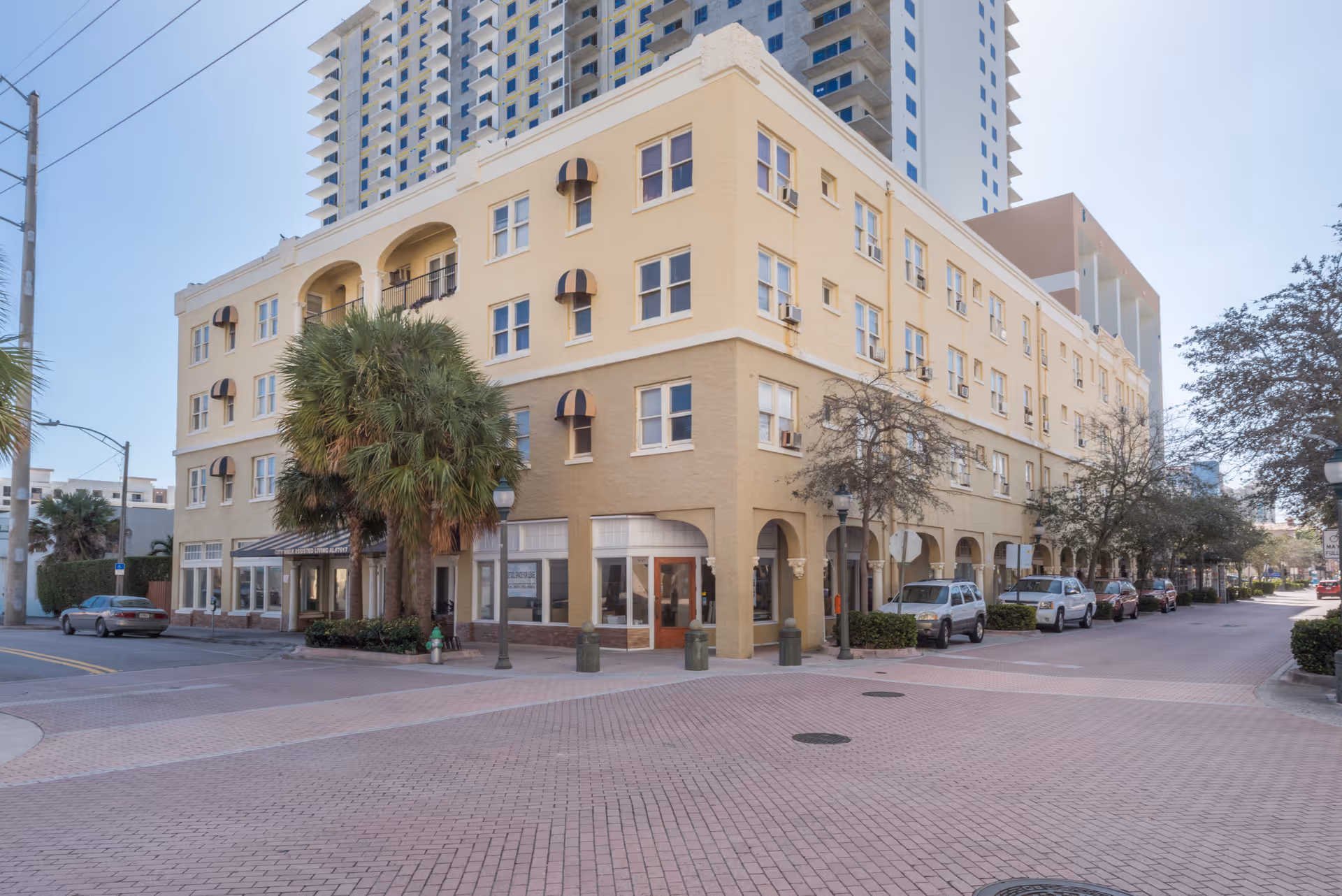 Exterior view of a multi-story beige building with arched windows and awnings, located on a corner with palm trees and parked cars along the street.