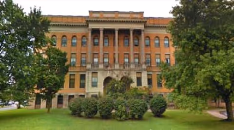 Front view of a large historic multi-story brick building with tall columns, surrounded by trees and a grassy lawn.