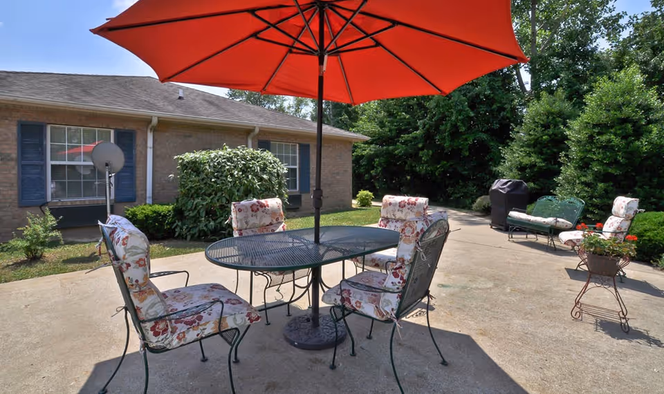 Outdoor patio area with a round metal table and four cushioned chairs featuring floral patterns. A large red umbrella provides shade over the table. In the background, there is a brick building with blue shutters and windows, surrounded by green bushes and trees. Additional seating and a covered grill are visible on the concrete patio.