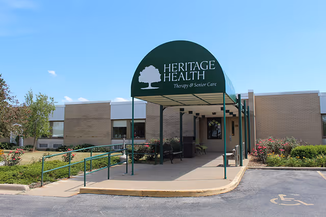 Exterior view of a single-story brick building with a green awning that reads 'Heritage Health Therapy & Senior Care'. There is a wheelchair accessible ramp and a parking space with a handicap symbol in front of the entrance. The area is landscaped with bushes and small trees under a clear blue sky.
