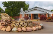 Exterior view of Cedar Ridge Health & Rehab Center featuring a brick building entrance with a peaked roof, a water fountain surrounded by stacked stone and large rocks in the foreground, and trees and shrubs around the area.