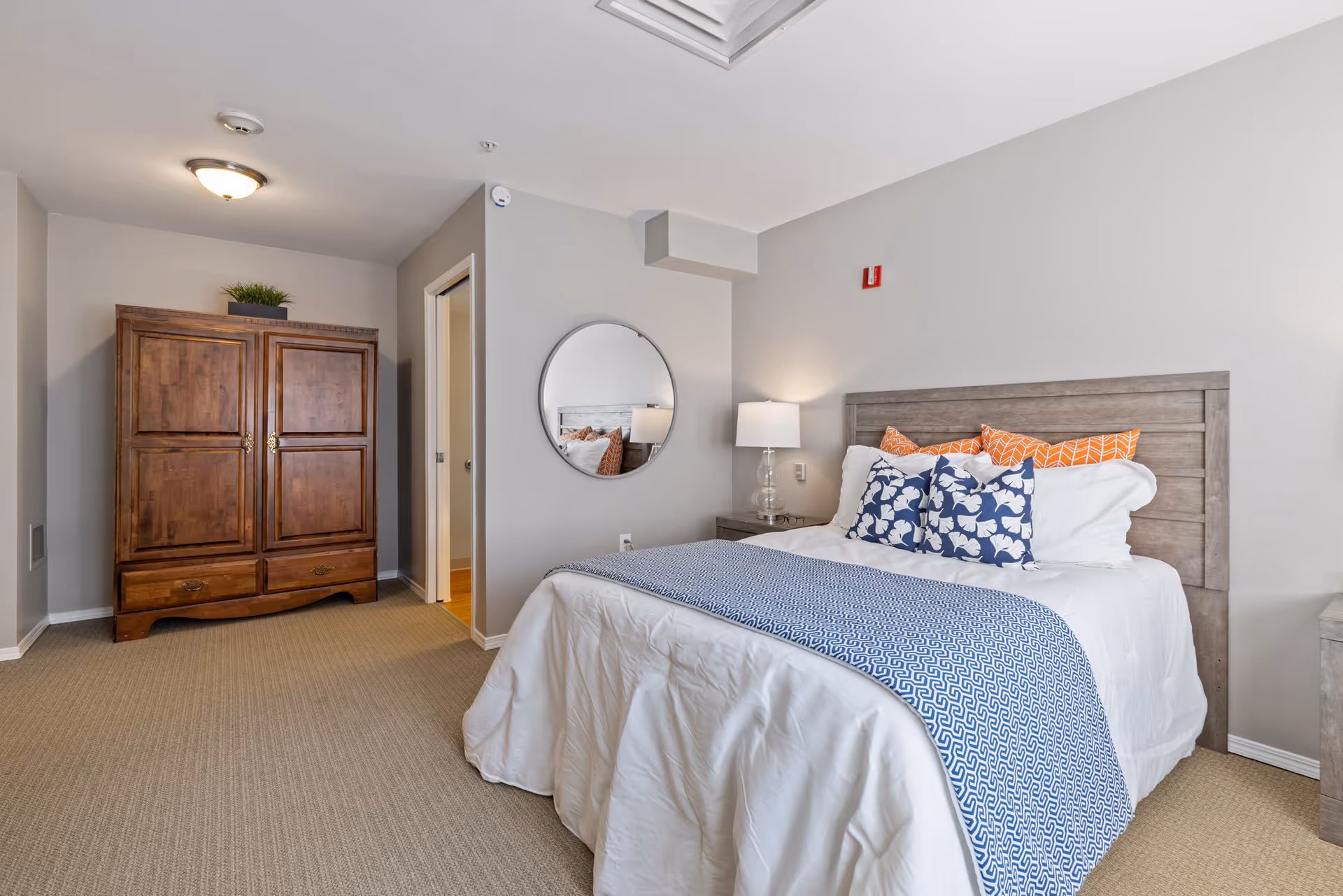 A bedroom with a neatly made bed featuring white bedding, a blue patterned throw, and decorative pillows in blue and orange. There is a wooden headboard, a nightstand with a lamp, a round mirror on the wall, and a wooden armoire with a plant on top. The room has beige carpet and light gray walls.