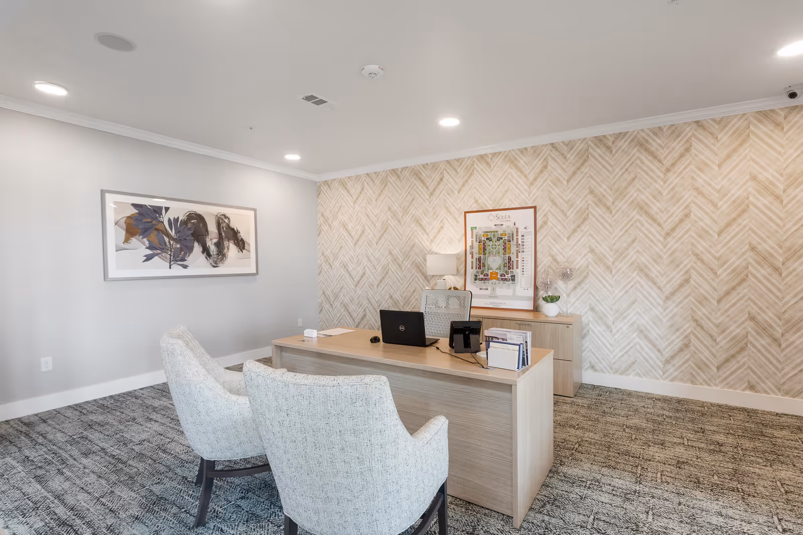 A modern office space with a wooden desk, two upholstered chairs, a laptop, a phone, and some documents. The room has patterned carpet flooring, a textured beige wall with a framed map labeled Solea Cinco Ranch, a table lamp, and a framed abstract artwork on the adjacent wall.