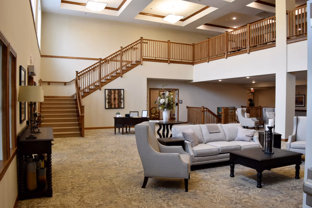 Spacious senior living facility common area with beige walls and carpet. The room features a light gray sofa, armchairs, a dark wooden coffee table with candles, and a round table with a large floral arrangement. There is a wooden staircase with railings leading to an upper level balcony. A desk with a computer and framed pictures is positioned against the far wall.