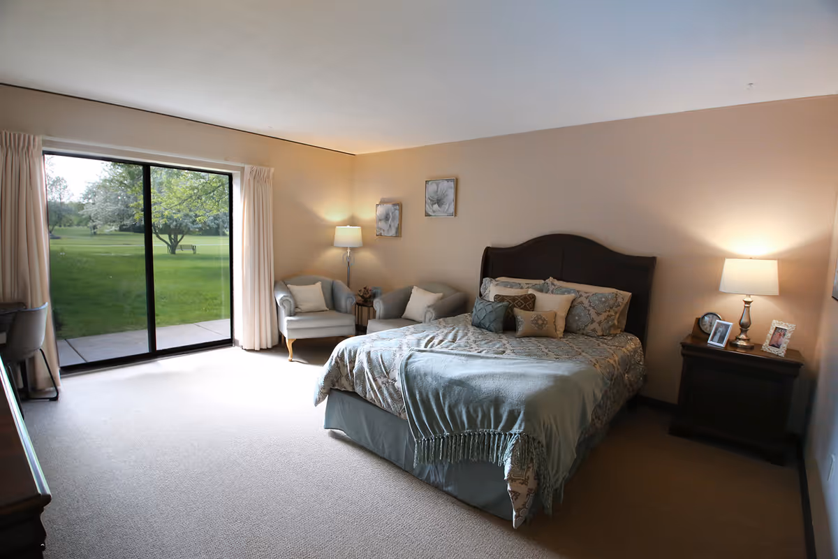 Well-lit furnished bedroom with a large bed, nightstands, seating area and sliding glass doors opening to a green lawn.