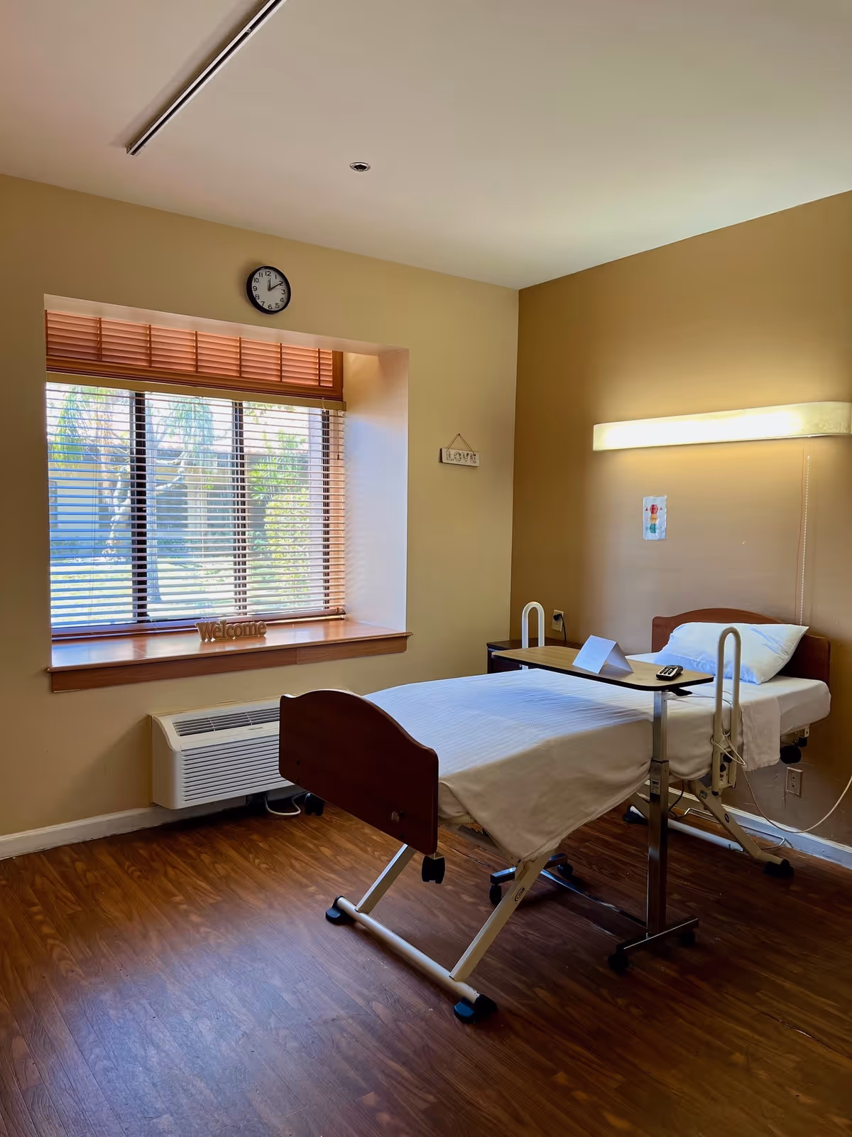 A simple, clean room with a single hospital-style bed covered with white sheets and a pillow. There is a small overbed table with a folded paper and a remote control on it. The room has wooden flooring, beige walls, a large window with wooden blinds, and a wall-mounted light above the bed. A clock is mounted above the window, and a small decorative sign that says 'welcome' is placed on the window sill.