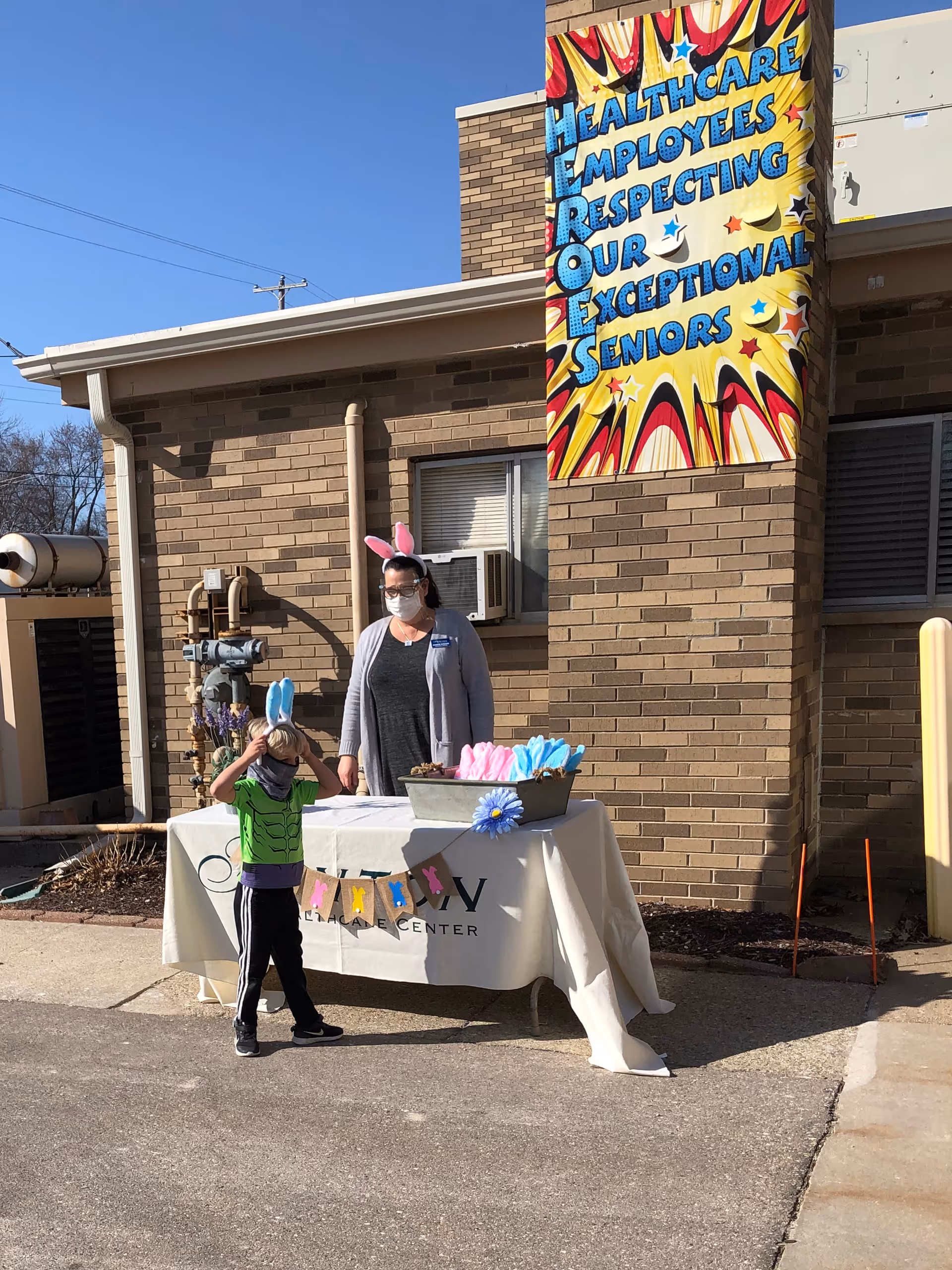 A woman wearing a face mask and pink bunny ears stands behind a table covered with a white cloth outside a brick building. A young child wearing a green shirt and blue bunny ears stands in front of the table. The table has a banner and containers with pink and blue items. A colorful sign on the building reads 'Healthcare Employees Respecting Our Exceptional Seniors.'