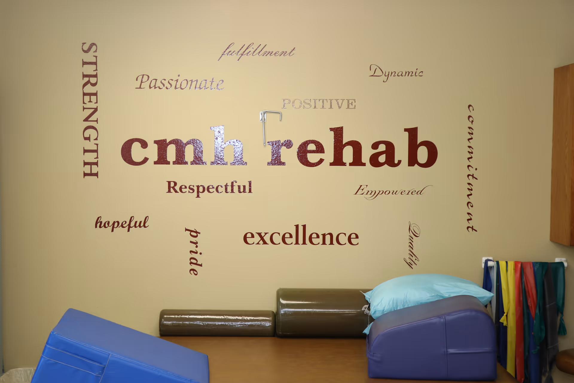 Therapy room interior showing a wall decal reading "cmh rehab" above exercise cushions and colorful resistance bands.