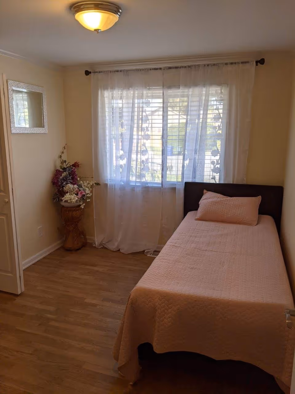 A small bedroom with a single bed covered in a light pink quilt and matching pillow. There is a window with sheer white curtains allowing natural light to enter the room. A decorative vase with artificial flowers is placed on a wooden stand in the corner, and a rectangular mirror hangs on the wall. The floor is wooden, and the walls are painted a light beige color.