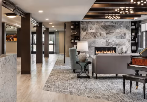 A modern and elegant living room area with a large stone fireplace, a gray sofa, a light blue armchair, a side table with a lamp, and a grand piano. The room features a patterned carpet, wooden ceiling beams with recessed lighting, and large windows allowing natural light to enter.