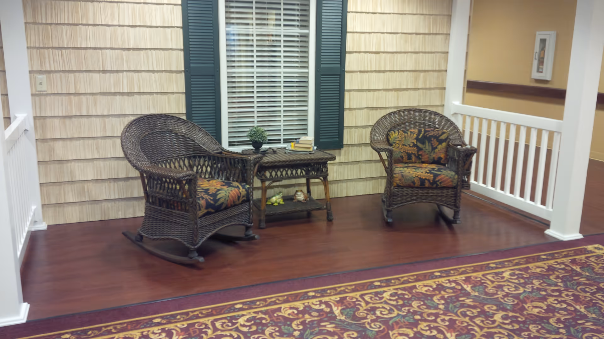 Indoor seating area with two wicker chairs featuring floral cushions and a small wicker table between them, placed on a wooden floor next to a window with green shutters and white blinds. The area is bordered by white railings and a patterned carpet is visible in the foreground.