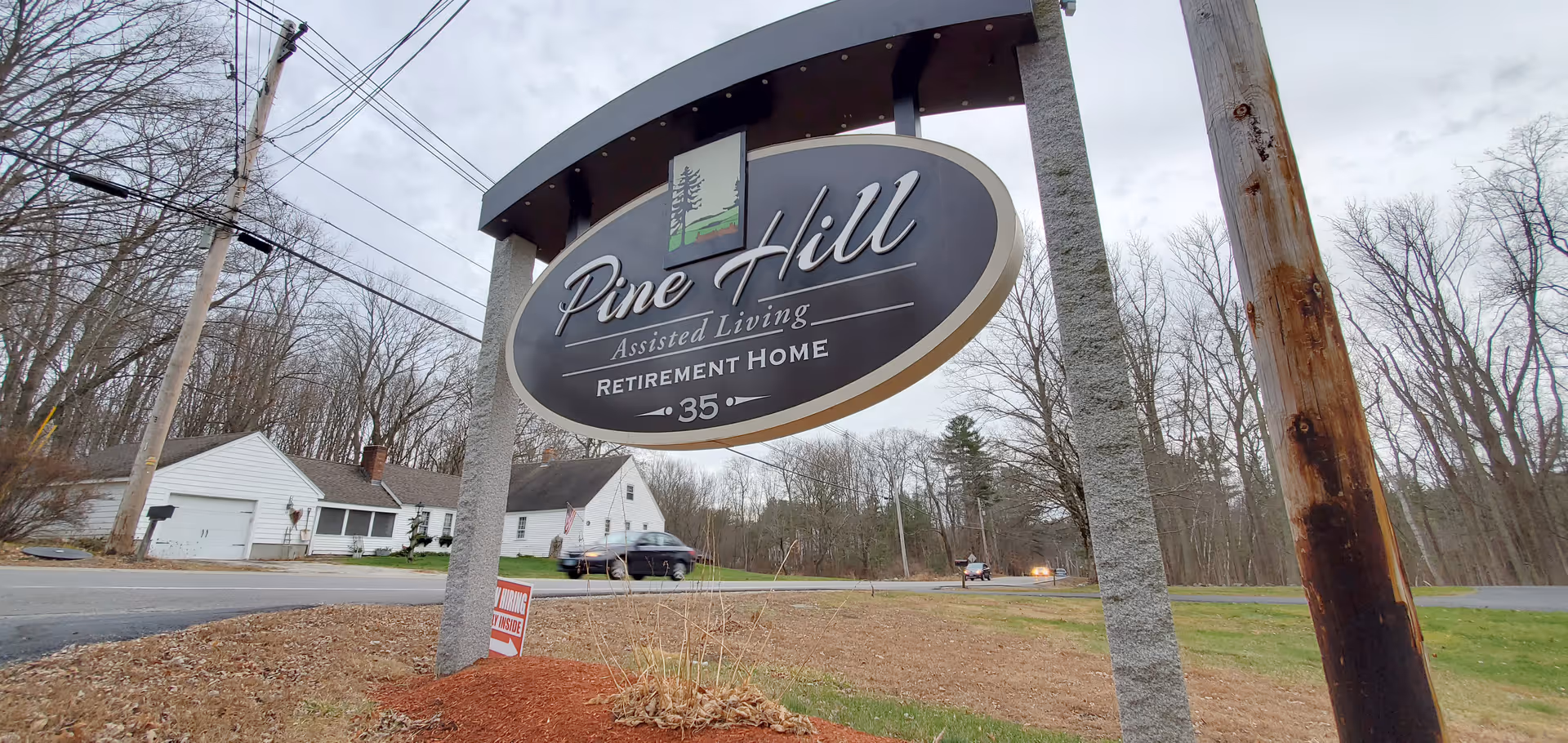Outdoor view of a large sign for Pine Hill Assisted Living Retirement Home with the number 35, situated near a road with houses and trees in the background under a cloudy sky.