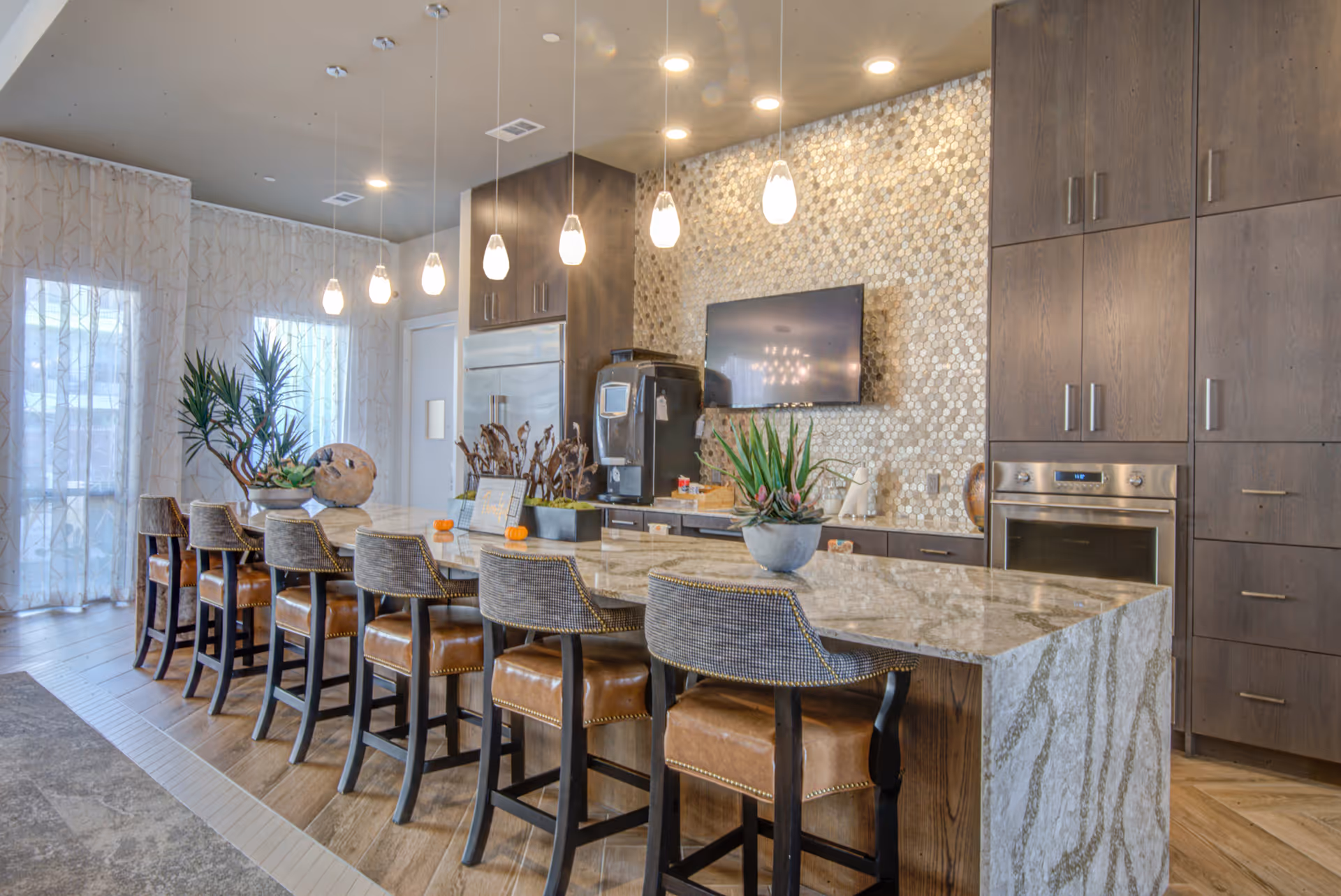 Modern kitchen area with a long marble island countertop surrounded by eight high-back chairs with leather seats. The back wall features a textured tile backsplash, a mounted flat-screen TV, and dark wood cabinetry with built-in stainless steel appliances. Pendant lights hang above the island, and large windows with sheer curtains allow natural light to fill the space. Decorative plants and small pumpkins are placed on the countertop.