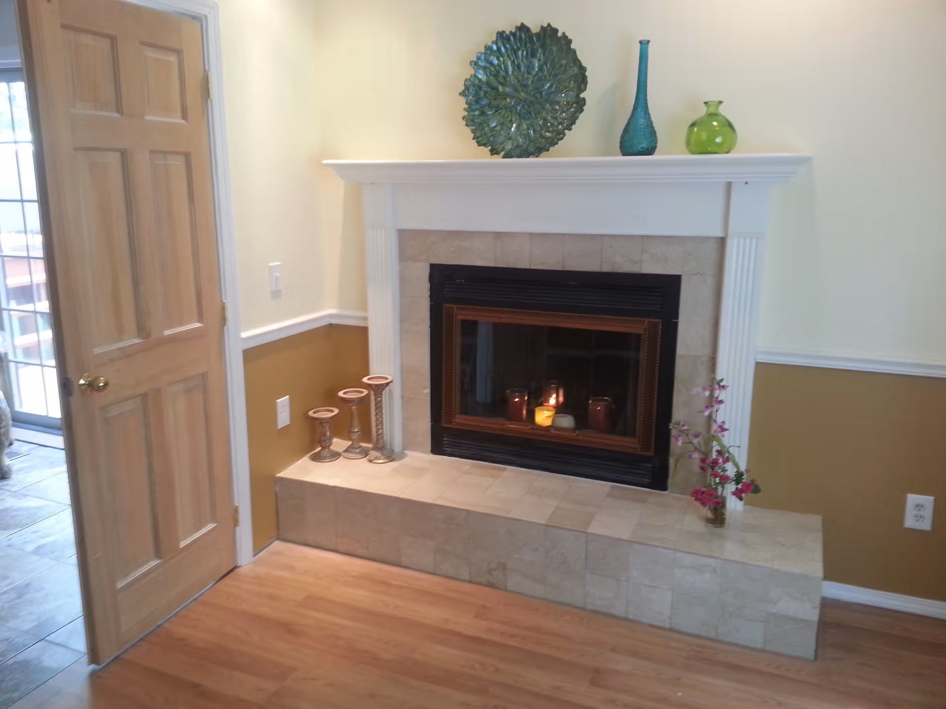 A living room fireplace with a white mantel topped by decorative vases and candles, beside a wooden door.