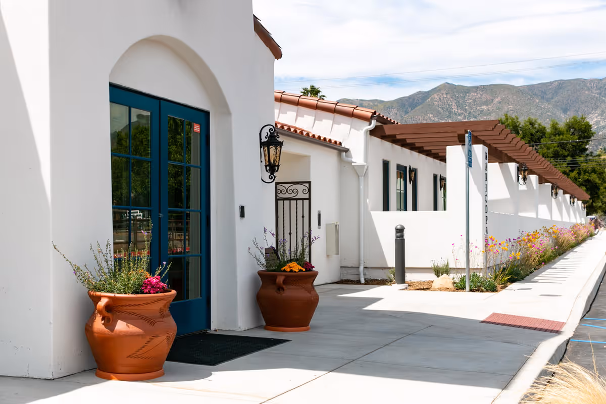 Exterior view of The Artesian of Ojai facility showing white stucco walls, blue double doors, terracotta roof tiles, large terracotta planters with flowers, a sidewalk, and mountains in the background under a partly cloudy sky.