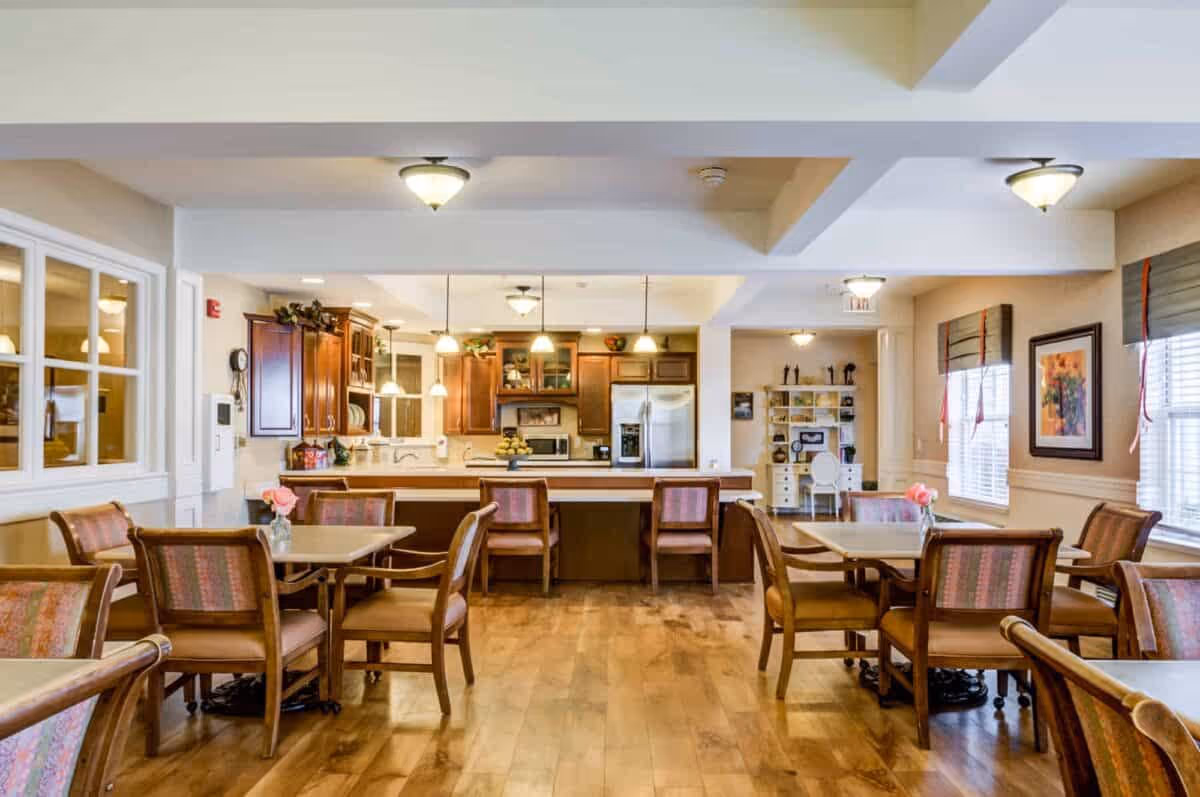 A bright and spacious dining area with wooden floors, multiple tables and chairs, and a kitchen in the background featuring wooden cabinets, a refrigerator, and pendant lighting. The room has large windows with blinds and framed artwork on the walls.
