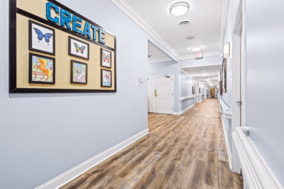 A long, well-lit hallway with light blue walls and wood-patterned flooring in a senior living facility. On the left wall, there is a framed display with the word 'CREATE' and colorful artwork including butterflies, a horse, a koala, and other images. The hallway has white doors and ceiling lights, with handrails along the walls.