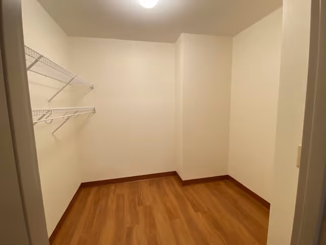 Empty room with wood flooring, white walls, and a white wire shelf mounted on the left wall under a ceiling light.
